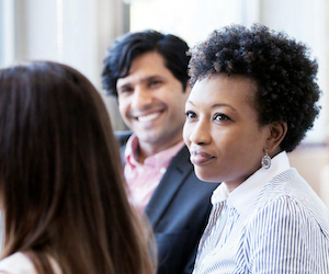 A woman wearing a blouse looks at someone with long hair as a man with a suit jacket in the background smiles broadly.